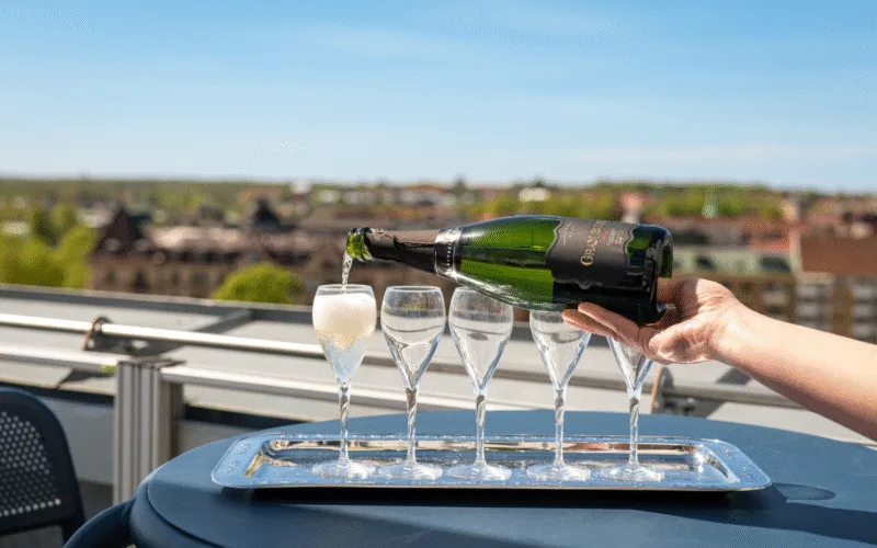 En person häller upp mousserande vin i ett glas på en bricka med flera glas på en terrass med utsikt över staden.