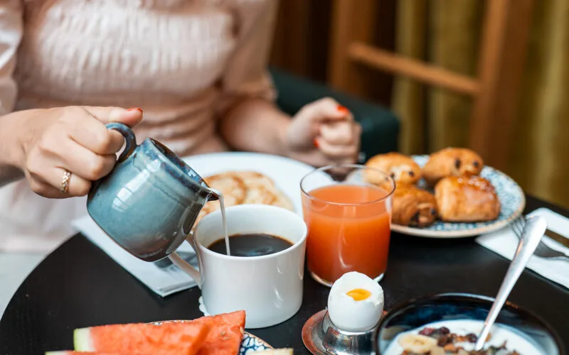 A person pouring milk into coffee at a breakfast table with juice, eggs, fruit, and pastries.