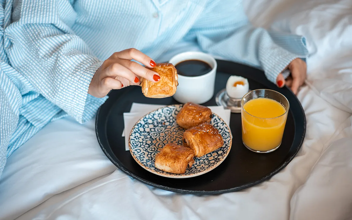A person in a bathrobe enjoys breakfast in bed with coffee, juice, a boiled egg, and freshly baked croissants on a tray.