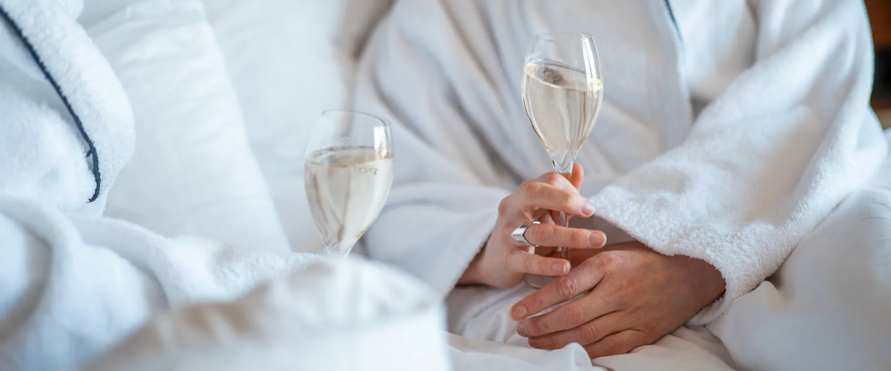 Två personer sitter i morgonrockar på en säng och skålar med bubbel