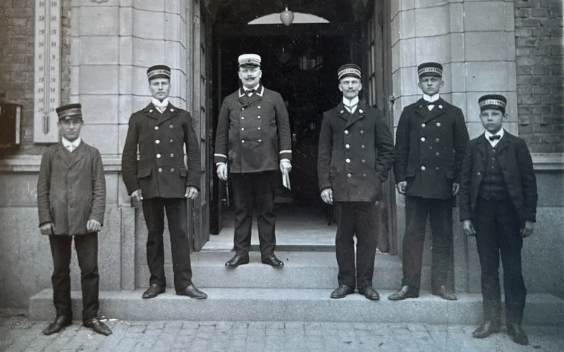Ett historiskt svartvitt foto med personal ståendes på Calmar Stadshotells trappa iklädda hatt och kostym.